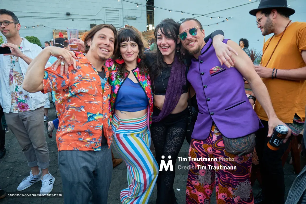 A jubilant group of five friends embraces in the golden afternoon light at White Owl Social Club's rooftop venue in Portland, Oregon. Their eclectic vintage-inspired outfits create a kaleidoscope of color against the urban backdrop, while string lights overhead hint at the festive atmosphere of the Your Sunday Best season opener. The genuine laughter and tight embrace capture the euphoric spirit of summer day parties, as revelers dressed in Hawaiian shirts, rainbow stripes, and purple blazers toast the beginning of another season of outdoor celebrations.