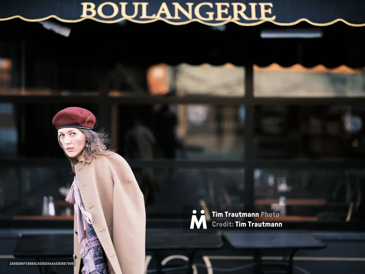 A young woman in a burgundy beret and camel coat glances back with an enigmatic expression as she passes a traditional French bakery storefront in Portland's Alberta Arts District. The warm golden lettering of 'BOULANGERIE' creates a romantic backdrop against the dark awning, while soft natural light illuminates her contemplative face. The composition captures a fleeting moment of urban European charm transplanted to the Pacific Northwest, with the subject's vintage-inspired styling echoing the old-world aesthetics of the bakery behind her.