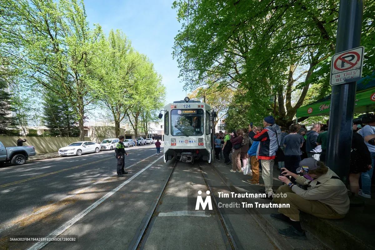 Enthusiasts gather along Northeast 11th Avenue in Portland's Lloyd District as TriMet's white Type 1 MAX train makes its ceremonial final run on April 18th, 2026. Dappled sunlight filters through mature street trees onto the crowd of transit fans and photographers positioned along the platform and trackside, creating a nostalgic atmosphere for this historic goodbye event. The scene captures the intersection of urban transit history and community celebration, with onlookers of all ages bearing witness to the end of an era in Portland's public transportation legacy.