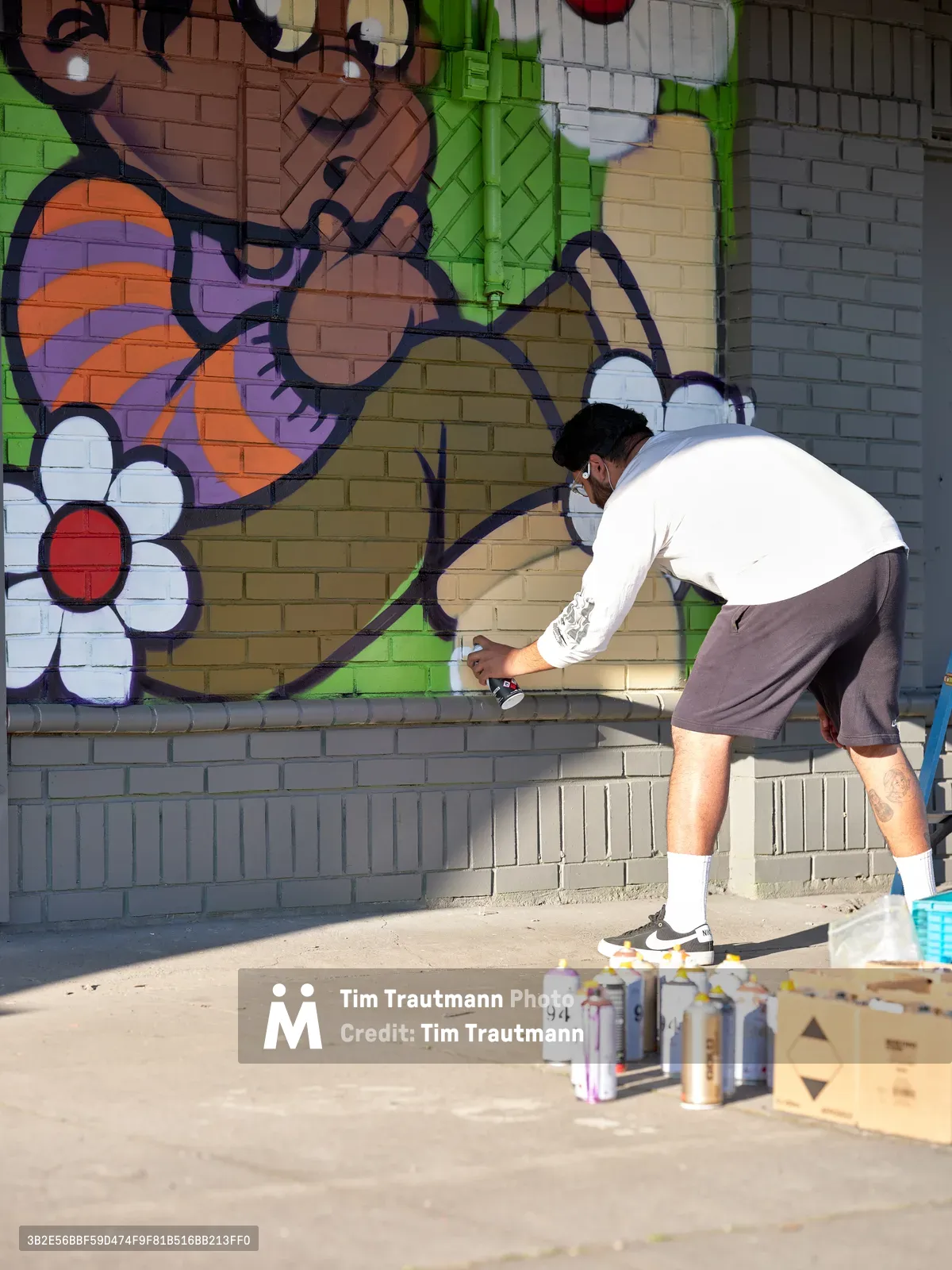 Muralist Heysus leans forward in concentrated effort, spray can in hand, adding intricate details to a colorful wall mural in Portland's Alberta Arts District. The artwork blooms across the brick surface with whimsical characters, bold florals, and a riot of greens, purples, and oranges. Scattered aerosol cans and a wooden crate of supplies rest on the sunlit sidewalk, evidence of the creative process unfolding in this corner of Northeast Portland where street art transforms urban landscapes.