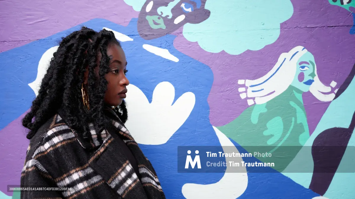 Artist Naomi Likayi sits in profile against her vibrant mural work at the boarded Portland World Trade Center, commissioned by the Portland Street Art Alliance. Her contemplative pose and striking locs create an intimate portrait against the dynamic backdrop of purple, blue, and mint green figures that seem to reach and interact across the wall. The interplay between the artist's earthtone plaid jacket and the bold street art creates a compelling juxtaposition of personal style and public expression.