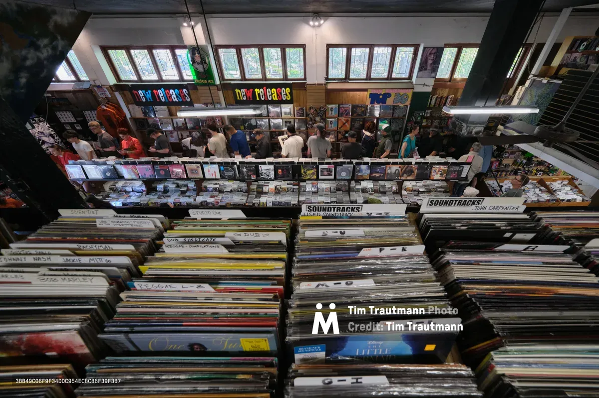 A bird's-eye view captures the bustling interior of Music Millennium record store on East Burnside Street in Portland's Kerns neighborhood. Towering stacks of vinyl records create geometric patterns in the foreground, while customers browse the extensive collection beneath warm fluorescent lighting. The store's signature colorful signage announces "New Releases" above orderly displays of albums, creating a nostalgic temple to analog music culture.