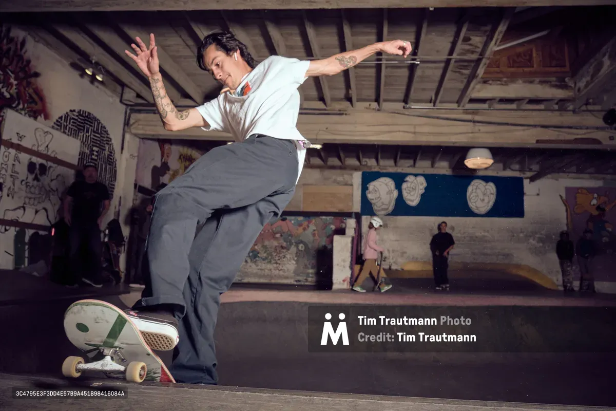 A tattooed skateboarder carves through Commonwealth Skateboarding's concrete bowl in Portland's Buckman neighborhood, his white shirt and grey pants contrasting against the graffiti-adorned walls. The underground skate park's raw industrial atmosphere comes alive with movement as spectators gather in the background beneath exposed ceiling beams. Warm tungsten lighting creates dramatic shadows across the weathered concrete surfaces, capturing the authentic energy of Portland's skateboarding culture.