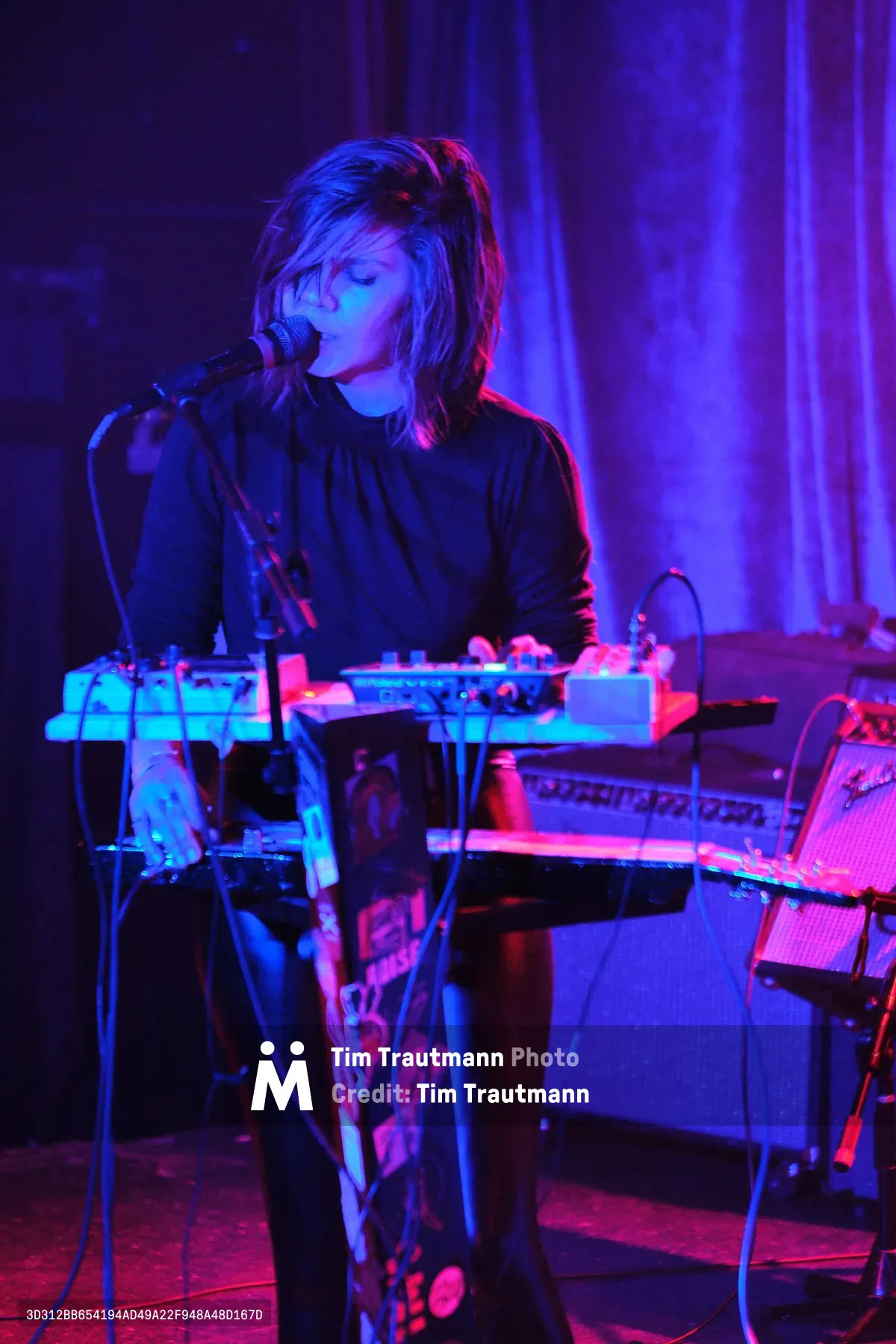 A solo electronic musician performs at Dante's in downtown Portland, bathed in ethereal purple and magenta stage lighting. The artist, wearing dark clothing, leans into a microphone while surrounded by synthesizers and electronic equipment that glow with cyan and pink hues. The intimate venue's curtained backdrop creates a moody atmosphere as the performer becomes one with their synthesized soundscape during this Rx Fest showcase.