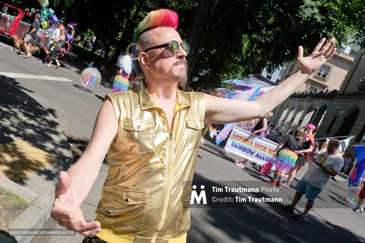 A charismatic parade participant in shimmering gold metallic attire commands attention with outstretched arms and rainbow-streaked hair beneath the dappled canopy of Portland's Pearl District trees. Soap bubbles drift through the sun-filtered air as colorful banners and enthusiastic crowds create a vibrant tapestry of celebration along Northwest 8th Avenue. The performer's theatrical pose and gleaming vest catch the filtered summer light, embodying the exuberant spirit of Pride festivities.