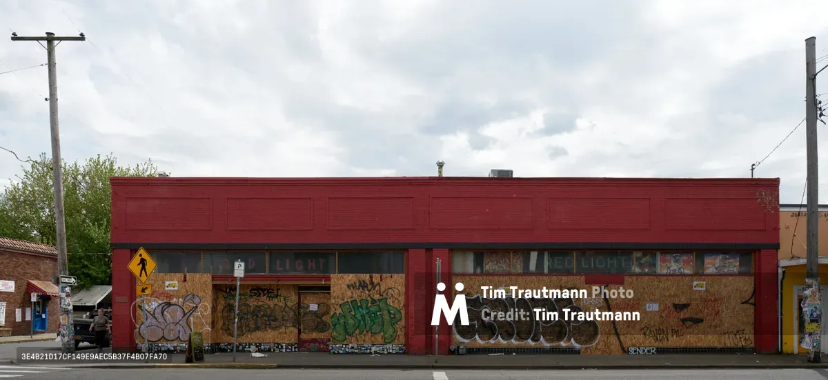 A weathered red brick commercial building stands shuttered along Southeast Hawthorne Boulevard in Portland's Richmond neighborhood, its windows and doors sealed behind plywood panels adorned with vibrant graffiti tags. The overcast Pacific Northwest sky casts a muted light across the vacant storefront, while utility poles and power lines frame the scene of urban abandonment. Colorful street art transforms the makeshift barriers into an impromptu canvas, creating a stark contrast between creative expression and economic closure.