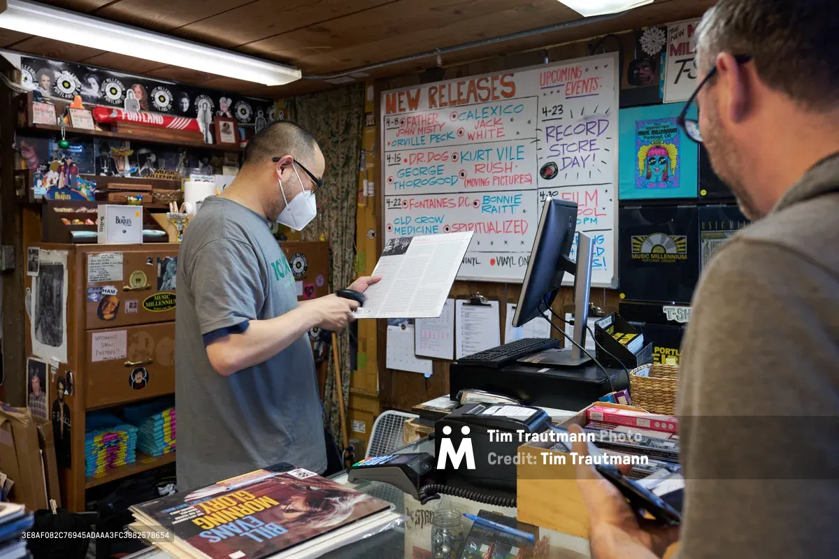 Inside the iconic Music Millennium record store on Portland's East Burnside Street during Record Store Day 2022, a masked employee in a gray t-shirt carefully reviews paperwork while a customer waits at the cluttered counter. The intimate shop buzzes with analog warmth, its wood-paneled walls plastered with vinyl records, concert posters, and a handwritten "New Releases" board listing upcoming albums. Fluorescent lighting casts an even glow over the transaction, capturing the enduring ritual of record store culture during this annual celebration of independent music retail in Oregon's music-loving city.