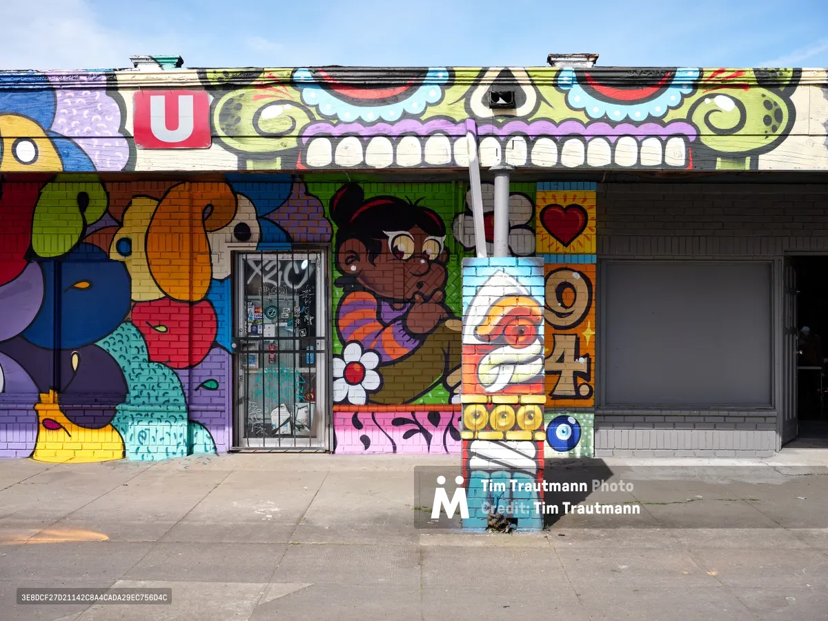 A kaleidoscopic street mural by artist Heysus 94 transforms the facade of Tuan's Auto Service in Portland's Alberta Arts District. The exuberant artwork features a menacing green skull with bared white teeth dominating the upper portion, while playful cartoon characters, abstract shapes, and bold typography cascade across the brick wall below. Bright afternoon light illuminates the saturated colors—electric blues, cherry reds, lime greens, and sunny yellows—creating a striking contrast against the weathered concrete sidewalk and clear blue sky.