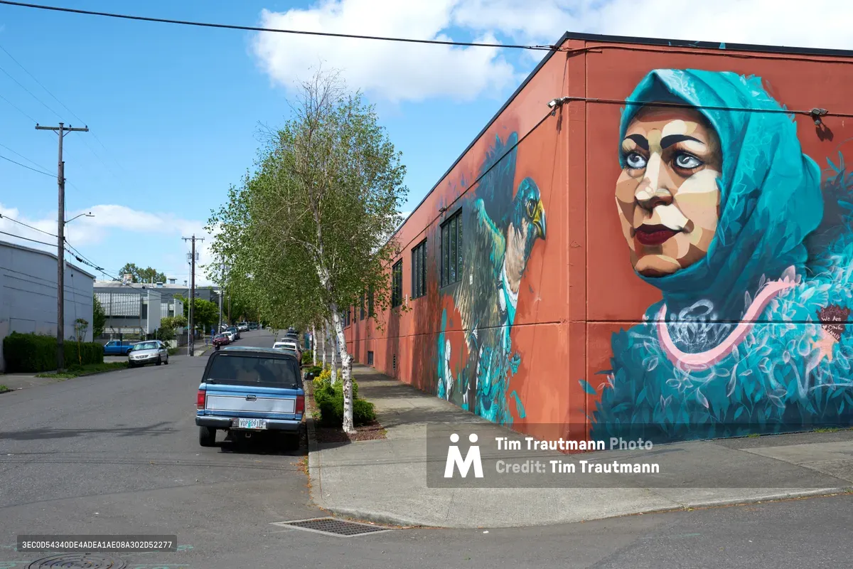 A monumental mural by artists Suhaib Attar and Ernesto Maranje transforms a rust-colored industrial building on Southeast Washington Street, depicting a woman with striking turquoise hair and penetrating gaze. The portrait emerges from flowing aquamarine brushstrokes against the building's weathered facade, while power lines and a leafy birch tree frame the scene under Oregon's characteristic cloudy sky. Part of Portland Street Art Alliance's community engagement initiative, this aptART-commissioned work brings vibrant artistic expression to the Central Eastside's industrial landscape.
