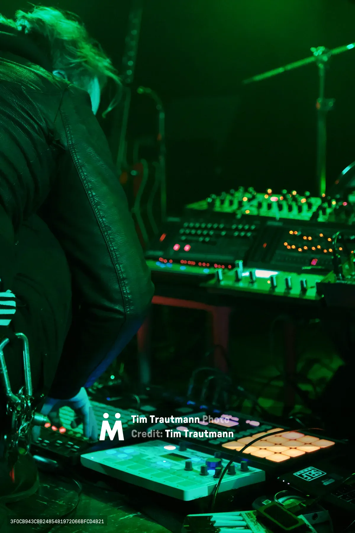 Electronic musician Eli Savage, performing as ellipsism, commands a complex array of synthesizers and electronic equipment bathed in ethereal green stage lighting at Portland's legendary Dante's venue. The intimate underground atmosphere captures the artist mid-performance, leather jacket and flowing hair silhouetted against the glowing constellation of buttons, knobs, and LED displays. Multiple levels of electronic instruments create a technological landscape while warm amber and cool green lights paint the scene in cyberpunk hues.