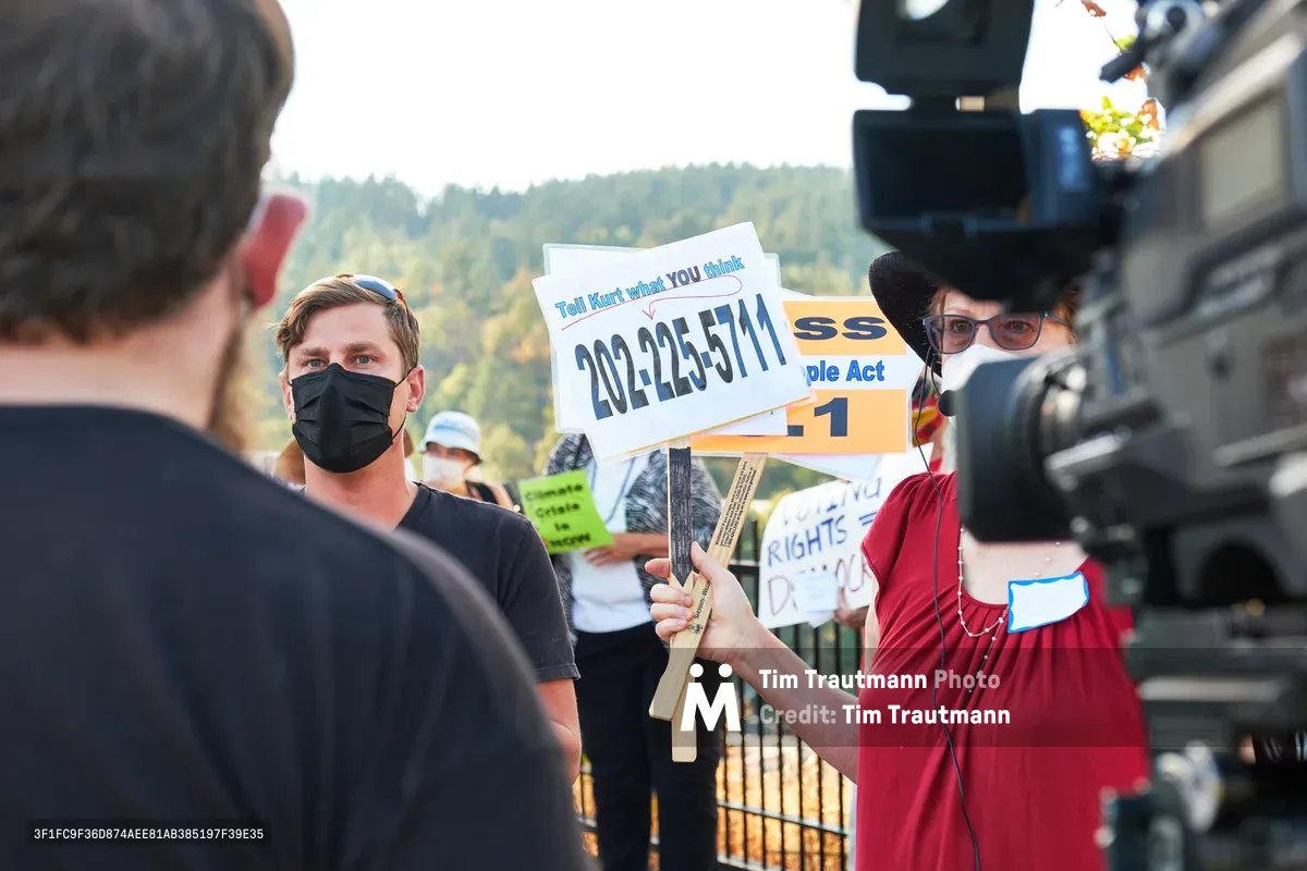 Michael Gaskill, wearing a black face mask, faces a television camera during a climate change protest outside Representative Kurt Schrader's office in Oregon City, Oregon. A demonstrator in period dress holds a protest sign displaying a phone number, while autumn-touched hills form a scenic backdrop. The scene captures the intersection of grassroots activism and media coverage, with bright afternoon light illuminating the passionate exchange between citizen and journalist.