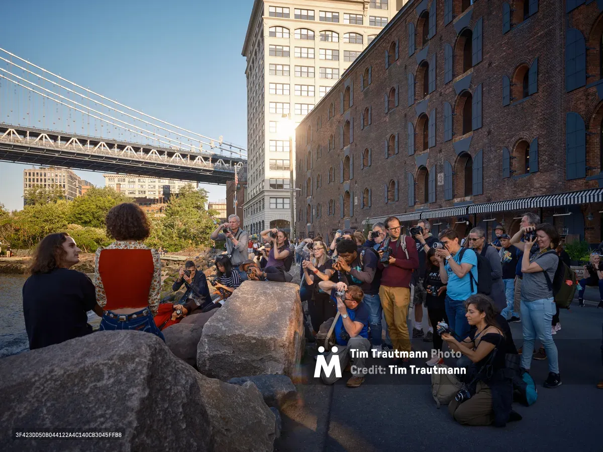 A vibrant gathering of photographers and onlookers assembles along the cobblestone waterfront beneath the Manhattan Bridge in Brooklyn's DUMBO neighborhood. The late afternoon light bathes the historic brick warehouses and modern high-rises in warm golden tones, while dozens of camera-wielding enthusiasts focus their lenses on subjects positioned among weathered granite boulders. The iconic suspension bridge cables stretch dramatically overhead, framing this quintessential New York photography scene where industrial heritage meets contemporary urban culture.