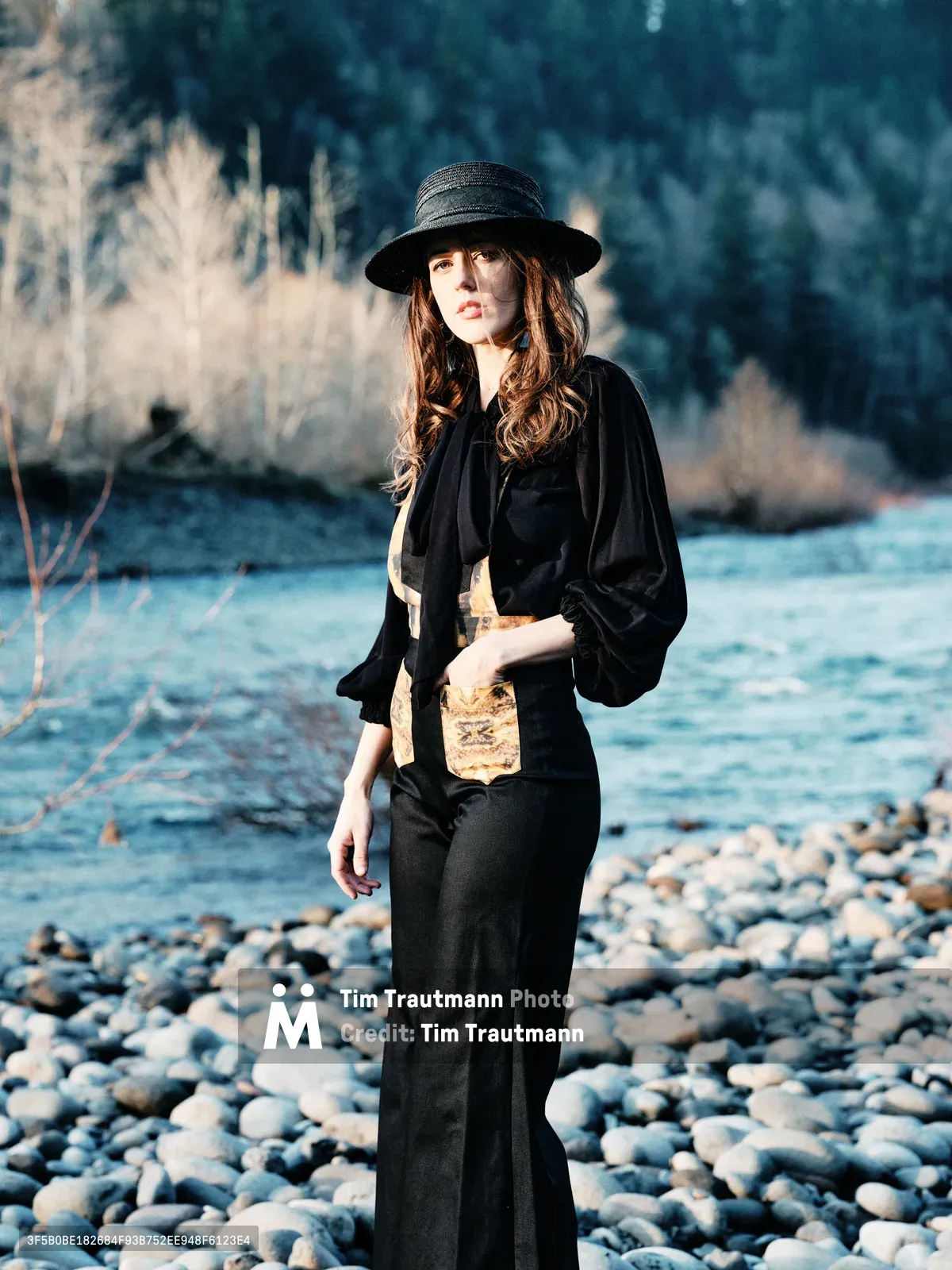 A contemplative woman in flowing black attire and wide-brimmed hat stands among the weathered river stones of Oregon's Floodplain Trail. Golden hour light illuminates her auburn hair and creates a dreamy atmosphere against the backdrop of dormant winter grasses and the flowing waters of Multnomah County's riparian landscape. The scene captures a moment of quiet reflection in the Pacific Northwest wilderness.
