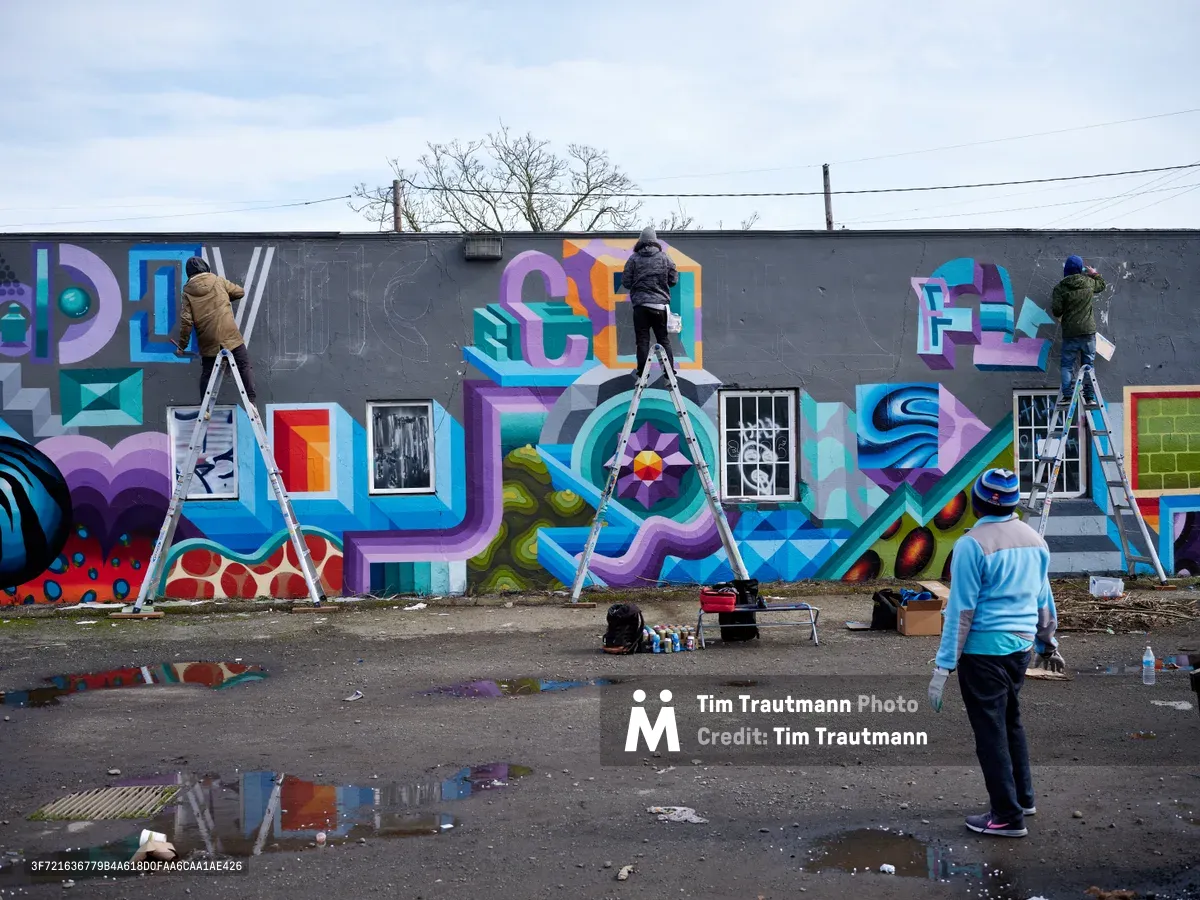 Multiple street artists work simultaneously on ladders to create a vibrant geometric mural along an industrial wall in Northeast Portland's arts district. The collaborative piece features bold dimensional letters spelling out words in blues, purples, and oranges against a charcoal background, while spray paint cans and art supplies scatter across the weathered asphalt below. The overcast Pacific Northwest sky provides soft, even lighting that illuminates the work-in-progress and the artists' focused concentration.