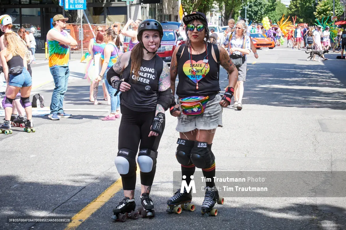 Two roller derby athletes pose confidently on Northwest 9th Avenue during Portland's 2019 Pride Parade, their protective gear gleaming in the bright midday sun. The left skater sports a 'Strong Athletic Queer' tank top while her teammate displays a rainbow heart necklace reading 'Skate Derby Hate,' both embodying the intersection of athleticism and LGBTQ+ pride. Behind them, the colorful parade unfolds through Portland's Pearl District, with rainbow-clad participants and spectators creating a vibrant tapestry of celebration along the tree-lined urban street.