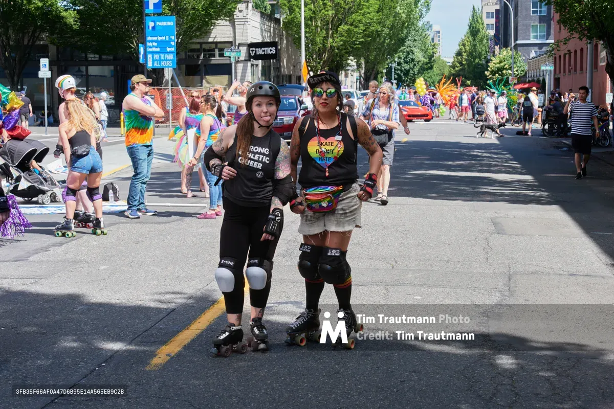 Two roller derby athletes pose confidently on Northwest 9th Avenue during Portland's 2019 Pride Parade, their protective gear gleaming in the bright midday sun. The left skater sports a 'Strong Athletic Queer' tank top while her teammate displays a rainbow heart necklace reading 'Skate Derby Hate,' both embodying the intersection of athleticism and LGBTQ+ pride. Behind them, the colorful parade unfolds through Portland's Pearl District, with rainbow-clad participants and spectators creating a vibrant tapestry of celebration along the tree-lined urban street.
