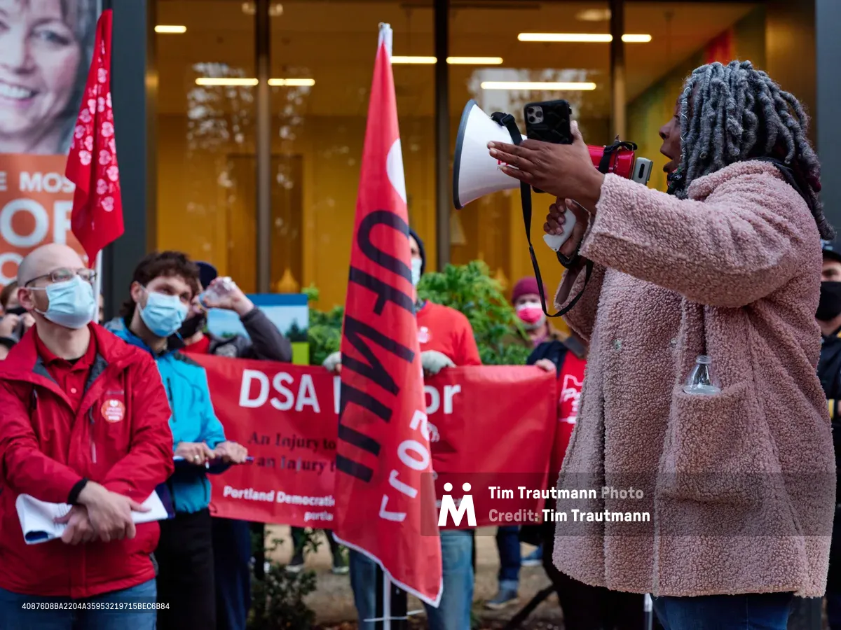 A passionate healthcare advocate with distinctive gray locs commands attention through a white megaphone, her voice cutting through the urban afternoon air outside Kaiser Permanente's imposing glass tower in Portland's Lloyd District. Behind her, masked nurses in crimson jackets clutch red DSA banners, their determined faces illuminated by the warm fluorescent glow spilling from the corporate lobby. The scene pulses with collective energy as workers prepare for their impending strike, their unified presence creating a stark contrast against the sterile corporate backdrop.
