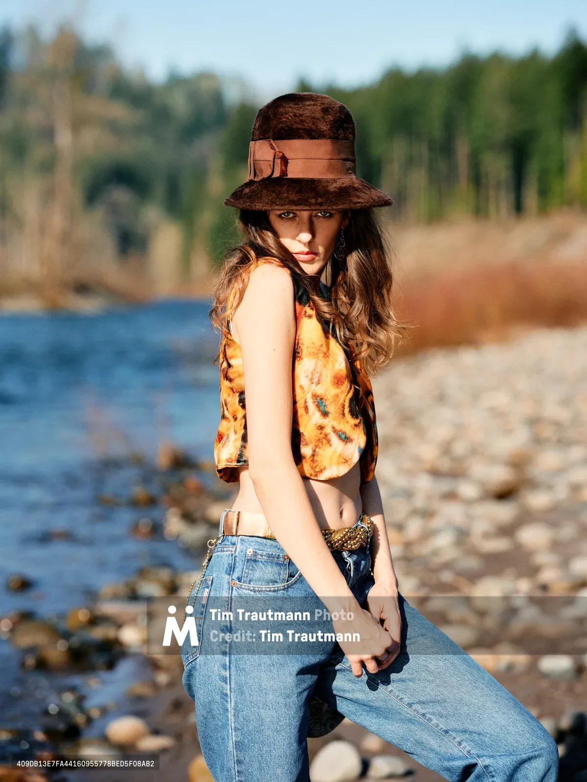 A young woman poses confidently along the rocky riverbed of Oregon's Floodplain Trail, her brown felt hat casting dramatic shadows across her face. She wears a vibrant tie-dye tank top in sunset hues paired with high-waisted denim, creating a striking contrast against the serene blue water and distant forest. The warm golden hour light bathes the scene, highlighting the natural textures of scattered stones and creating an intimate, contemplative mood.
