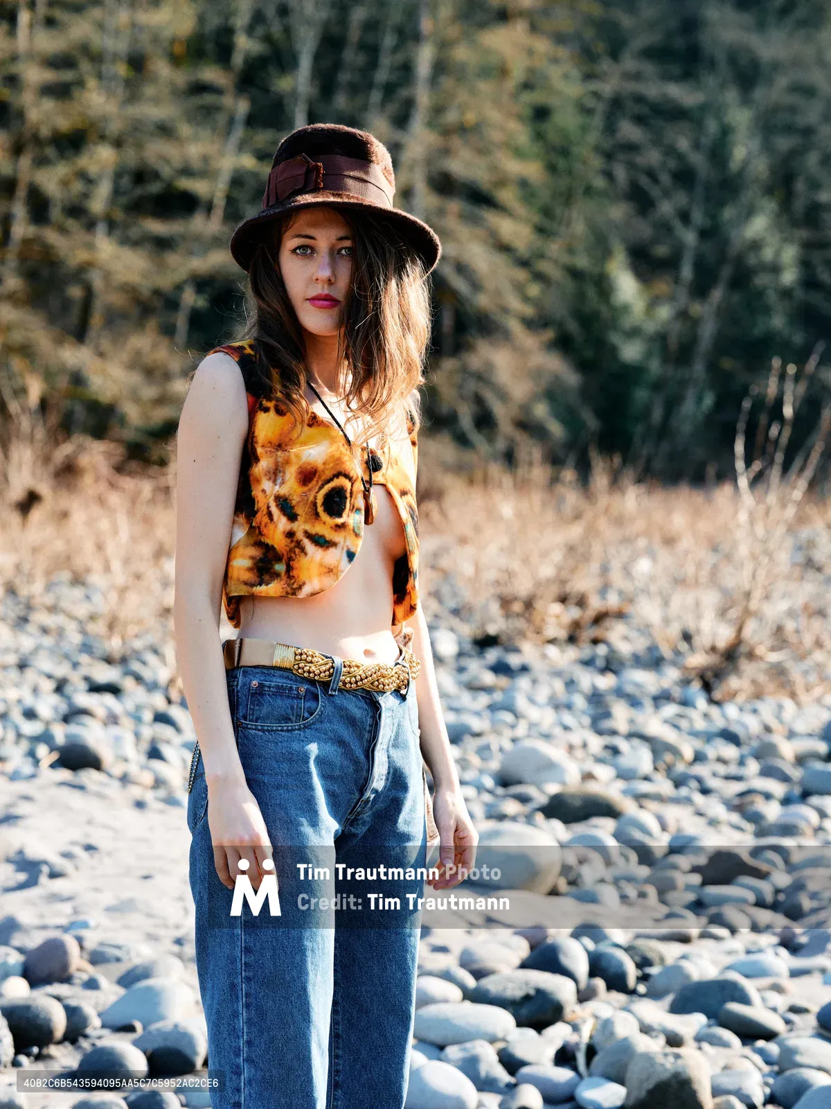 A young woman stands contemplatively among the sun-bleached stones of a dried riverbed along Oregon's Floodplain Trail. Golden afternoon light illuminates her vintage-inspired ensemble of high-waisted denim, a tie-dye crop top, and weathered leather hat, while autumn grasses and bare cottonwoods create a textured backdrop. Her direct gaze and relaxed stance suggest a moment of peaceful solitude in this riparian landscape near the Columbia River Gorge.