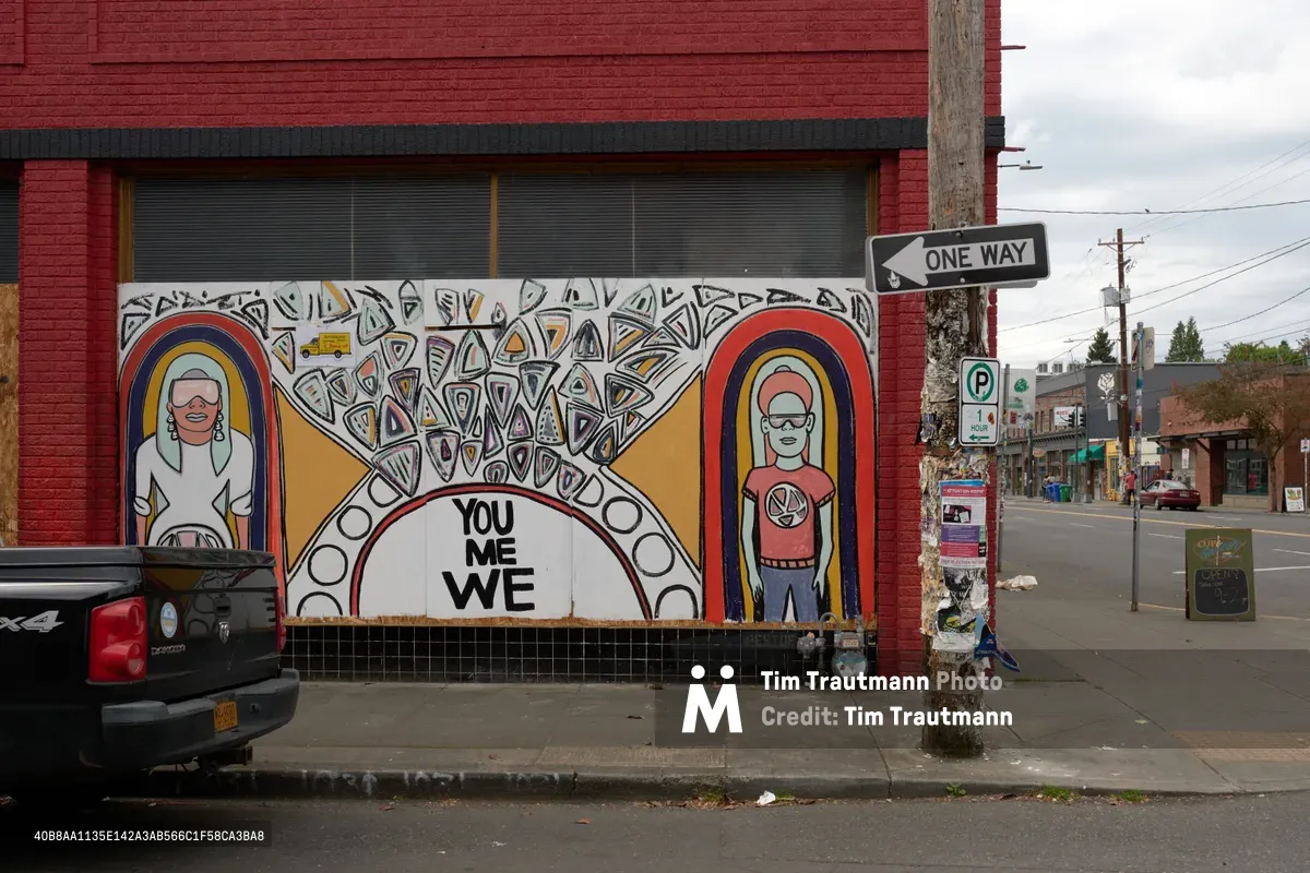 A vibrant community mural adorns the boarded windows of a closed business on Southeast Hawthorne Boulevard in Portland's Richmond neighborhood. The artwork features two portrait figures flanking a central mandala design with the message 'YOU ME WE' prominently displayed, transforming urban decay into a canvas for connection and hope. The red brick facade creates a striking backdrop against the overcast Pacific Northwest sky, while street signs and utility poles frame this intersection of abandonment and artistic resilience.