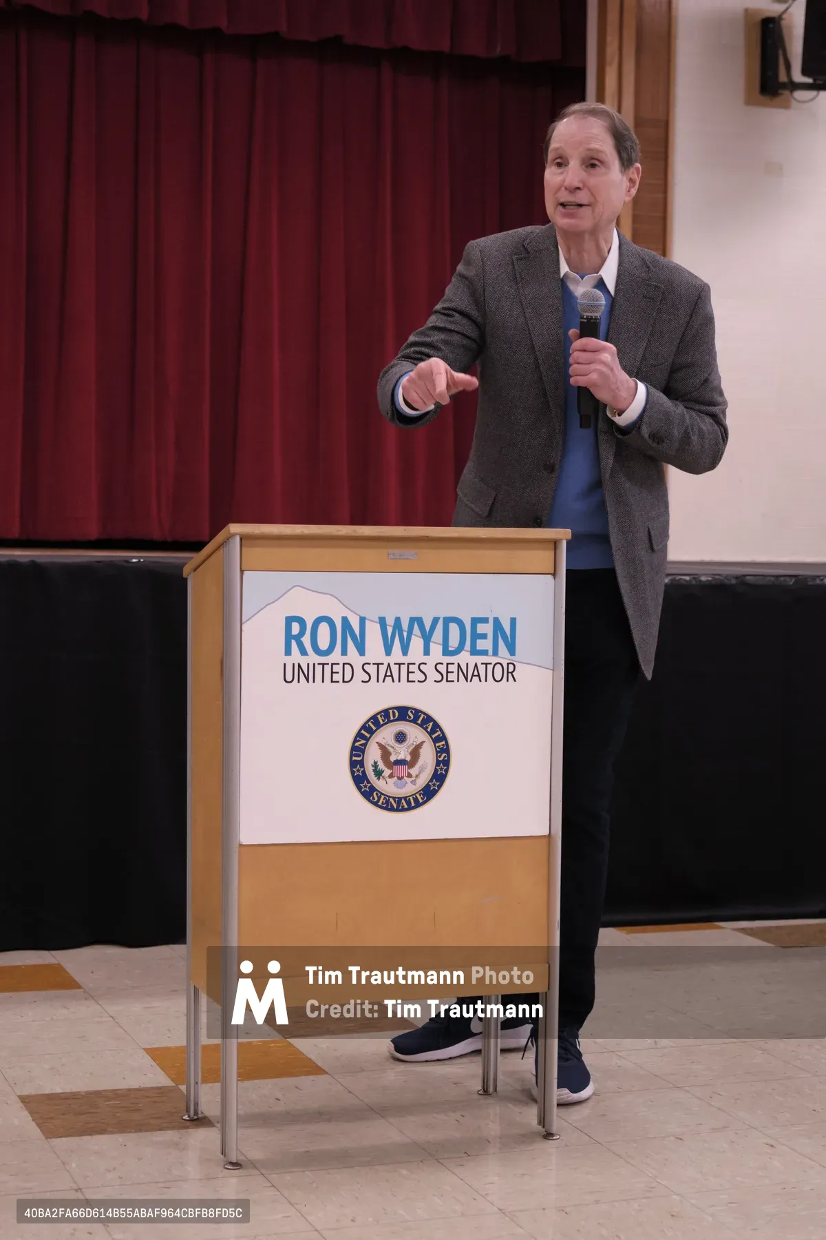 United States Senator Ron Wyden delivers remarks from behind a podium bearing his name and the Senate seal at Robert Gray Middle School in Portland's Hillsdale neighborhood. The seasoned politician, dressed in a charcoal blazer and blue shirt, gestures emphatically while speaking into a microphone, his expression animated against the backdrop of burgundy stage curtains. The institutional setting and formal podium setup capture the essence of grassroots democratic engagement in suburban Oregon.