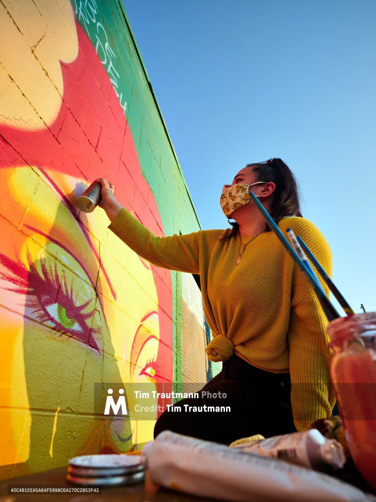 Multidisciplinary artist Vilatose works intently on her mural 'Jardín Cubana' depicting three female figures along Northeast Alberta Street in Portland's Alberta Arts District. Wearing a golden mustard sweater and patterned face mask, she applies paint with focused concentration against the backdrop of her emerging artwork's bold yellows, pinks, and greens. The bright afternoon light illuminates both artist and creation, capturing the dynamic energy of Portland's celebrated street art scene.