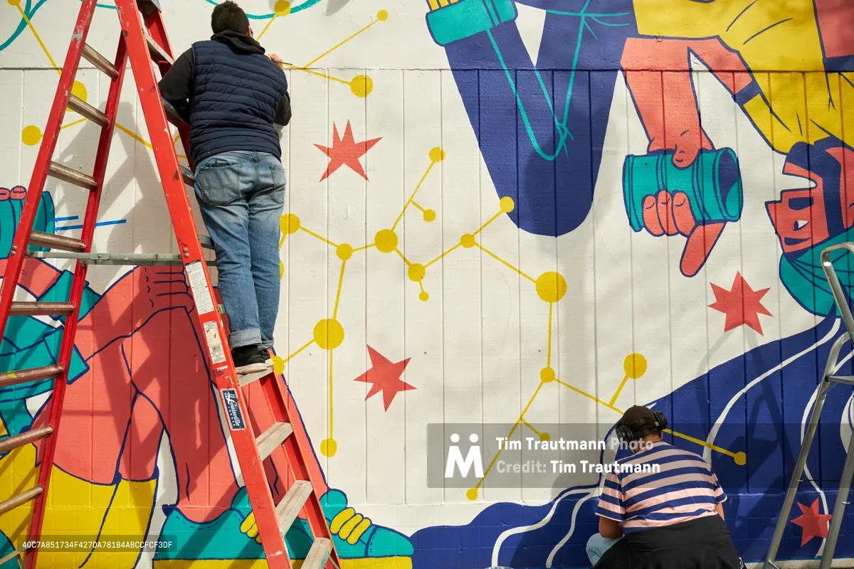 Two muralists work at different heights on a vibrant public art installation titled 'Let's Talk' at Open Signal in Portland's historic Eliot neighborhood. One artist climbs a red step ladder while another crouches near the base, both contributing to a dynamic composition featuring bold geometric figures, golden constellation-like networks, and scattered red stars against white concrete block walls. The collaborative scene captures the methodical process of large-scale community art creation in Northeast Portland's cultural corridor.