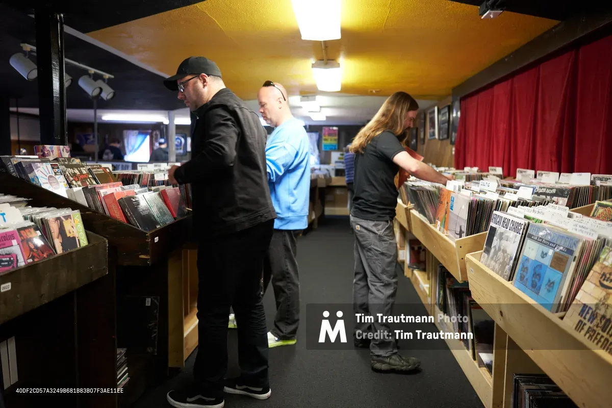 Three dedicated music enthusiasts browse through wooden crates of vinyl records at Portland's legendary Music Millennium record store on East Burnside during Record Store Day 2022. Warm overhead lighting bathes the intimate shop interior as a bearded man in black, a customer in blue, and a long-haired woman methodically flip through albums, each absorbed in their personal quest for musical treasures. The cramped quarters filled with organized chaos of record bins create an authentic atmosphere of analog discovery during this annual celebration of independent record stores.