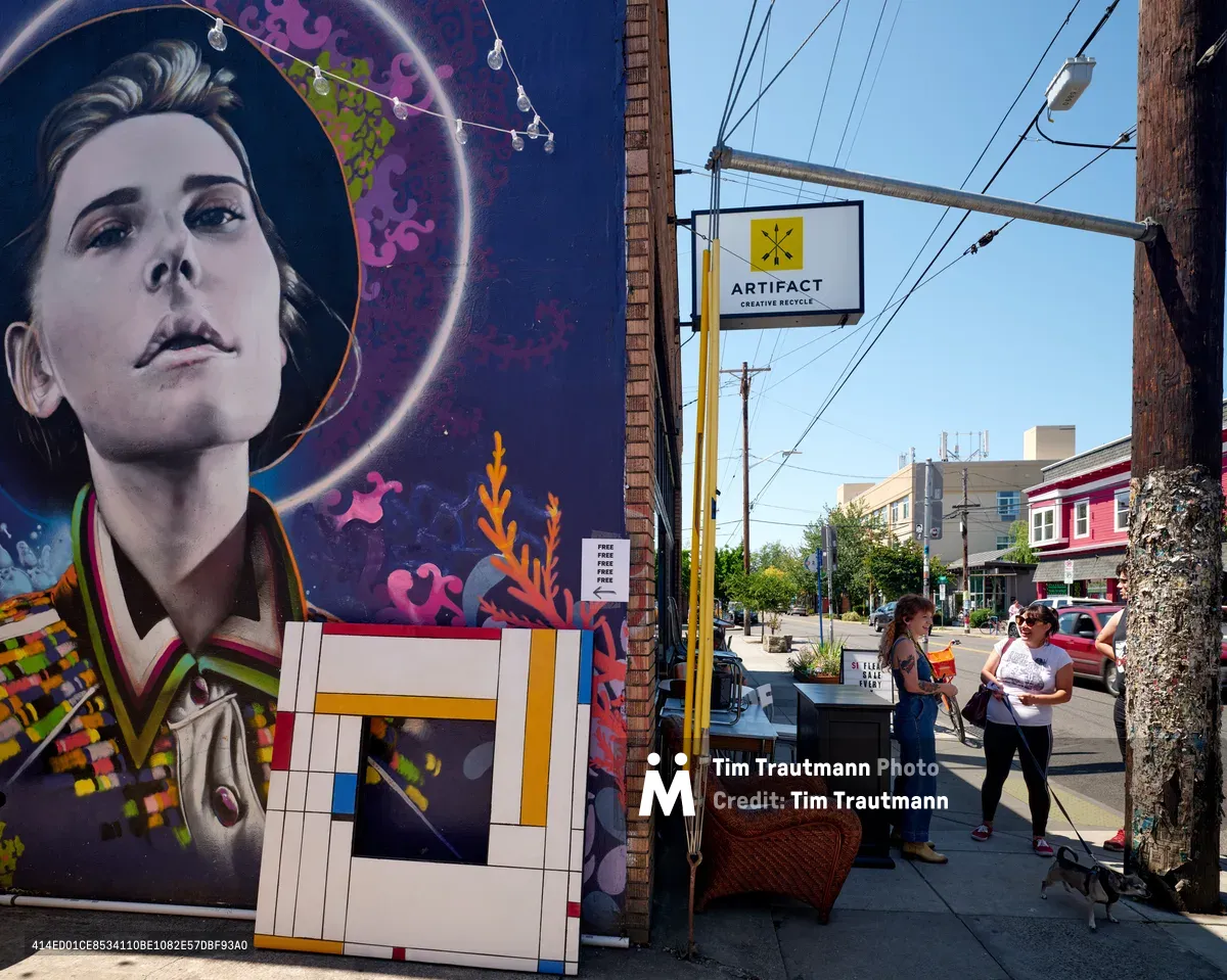 Two women engage in animated conversation outside Artifact Creative Recycle on Southeast Division Street, their casual interaction unfolding against a backdrop of striking street art. A monumental mural dominates the brick wall, featuring a portrait of a woman surrounded by cosmic swirls of deep purple and pink, complemented by geometric patterns reminiscent of Mondrian. The afternoon sunlight bathes the Richmond neighborhood scene, casting sharp shadows across the sidewalk where power lines create geometric patterns overhead.