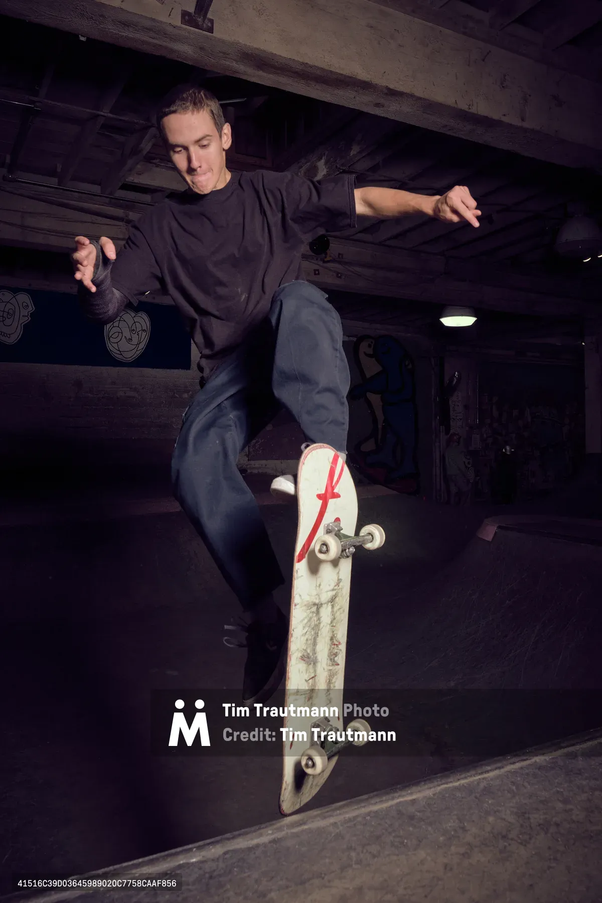A young skateboarder executes a fluid carve through the concrete bowl at Commonwealth Skateboarding in Portland's Buckman neighborhood. The moody lighting catches the weathered skateboard bearing a distinctive red 'F' logo as the rider maintains perfect balance through the curved transition. Exposed wooden beams overhead and the industrial atmosphere create an authentic underground skate culture scene, with the worn concrete and ambient shadows emphasizing the raw energy of street skating.
