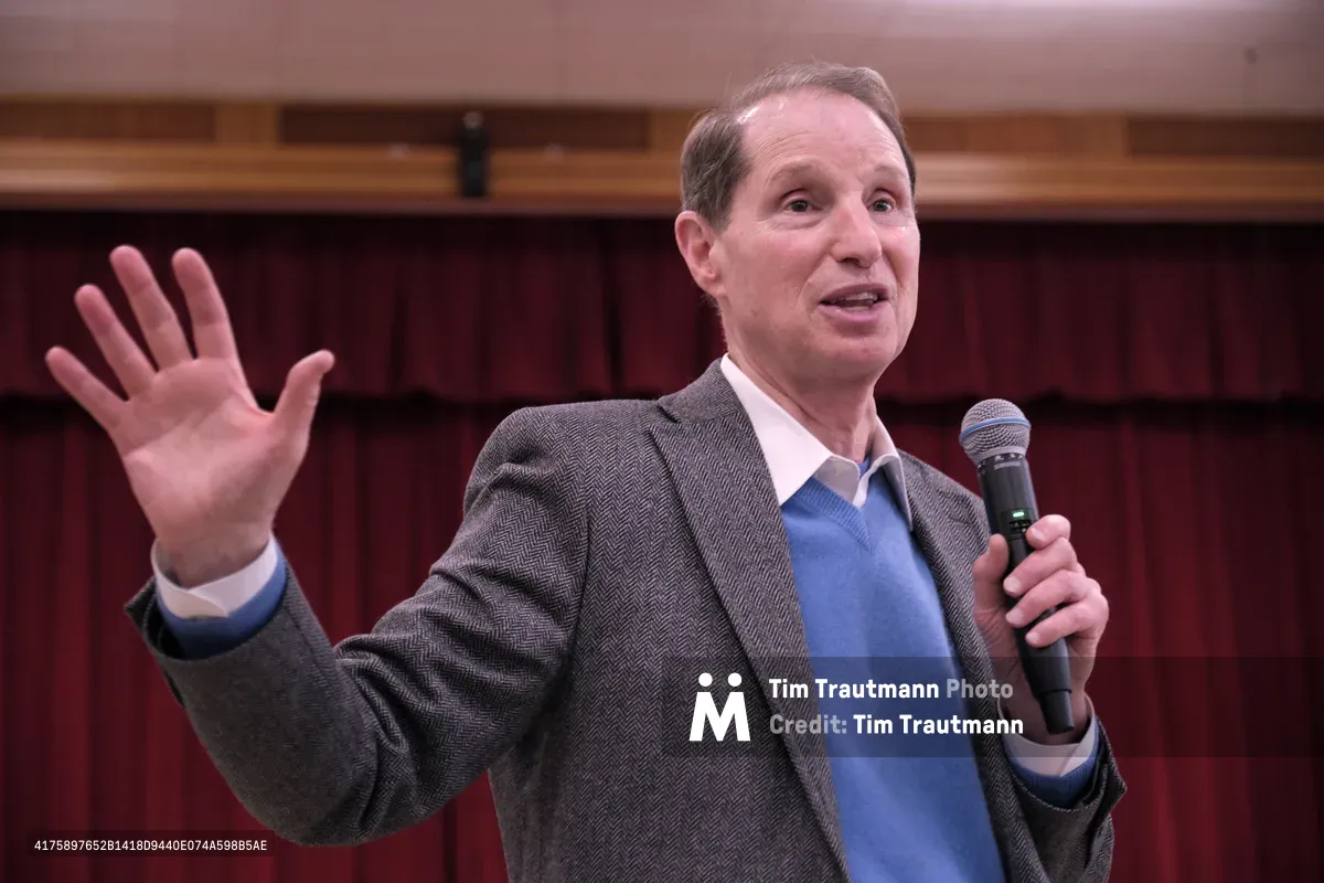 Oregon Senator Ron Wyden gestures emphatically while speaking into a handheld microphone during a town hall meeting at Robert Gray Middle School in Portland's Hillsdale neighborhood. The middle-aged politician, dressed in a herringbone sport coat over a blue sweater, raises his left hand in an expressive gesture as he addresses constituents. Behind him, the school's burgundy curtains and warm wood paneling create an intimate community gathering atmosphere.