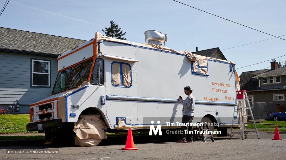 A muralist works diligently on Papa's Soul Food Kitchen Cart in Portland's King neighborhood, surrounded by residential houses and spring greenery. The artist, wearing a white long-sleeved shirt and baseball cap, stands beside the white food truck adorned with orange and blue trim, methodically applying paint while orange traffic cones mark the workspace. Afternoon light casts gentle shadows across the suburban street scene, capturing the quiet transformation of a neighborhood business through public art.