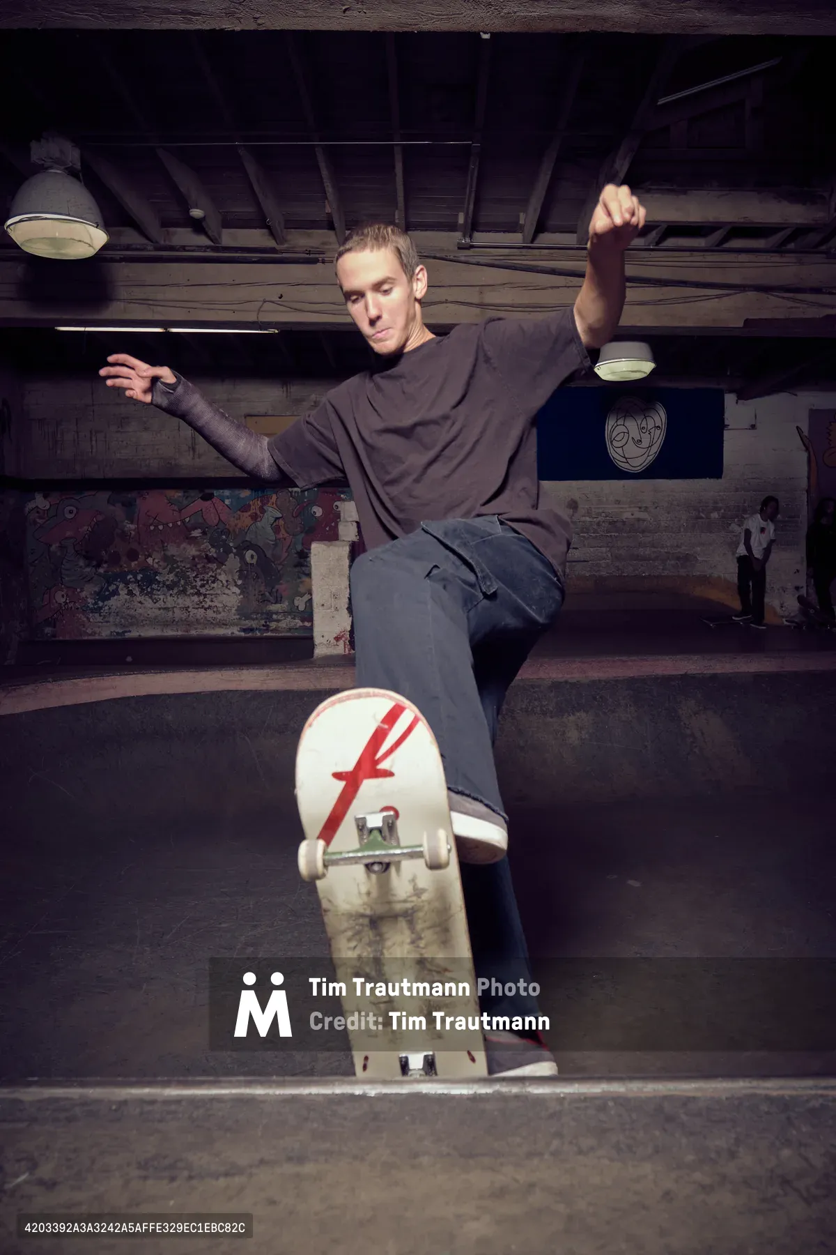 A focused skateboarder sits contemplatively on weathered concrete steps in the shadowy depths of Commonwealth Skateboarding in Portland's Buckman neighborhood. Arms outstretched in perfect balance, he embodies the zen-like concentration that defines bowl skating culture. The stark industrial lighting cuts through the underground atmosphere, illuminating weathered walls adorned with graffiti while his skateboard—marked with a distinctive red 'F' logo—rests against the worn concrete, telling stories of countless sessions in this subterranean sanctuary.