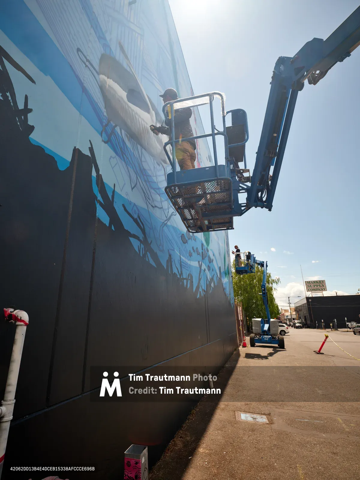 Jeremy Nichols and Devin Finley work from aerial lifts to create a large-scale mural on the concrete facade of a Southeast Portland building. The dramatic low-angle perspective captures the artists suspended against a bright sky as they paint flowing blue and black elements onto the industrial wall. The scene unfolds in Portland's Central Eastside district, where street art transforms urban landscapes through collaborative artistic endeavors supported by the Portland Street Art Alliance.