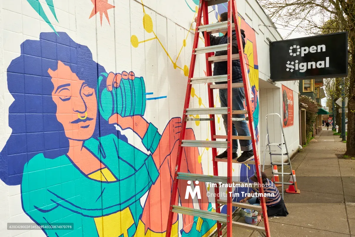 Two muralists work on a vibrant public art piece titled "Let's Talk" on the exterior wall of Open Signal community media center on Martin Luther King Jr. Boulevard in Portland's Eliot neighborhood. One artist perches on a red stepladder while painting detailed sections of the colorful mural, which features a figure with flowing blue hair holding turquoise objects against a backdrop of geometric patterns in coral, yellow, and teal. The scene captures the collaborative spirit of community art-making under overcast Pacific Northwest skies.