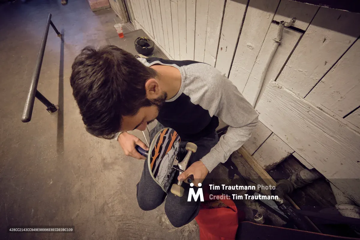 In the intimate confines of Commonwealth Skateboarding on Southeast 20th Avenue, a focused craftsperson kneels on weathered concrete, meticulously working on a custom skateboard deck. The scene unfolds beneath harsh fluorescent lighting that casts dramatic shadows across paint-chipped white wooden panels and scattered tools. Red equipment and materials punctuate the industrial workspace, while the artisan's concentrated posture speaks to the dedication required in board customization.