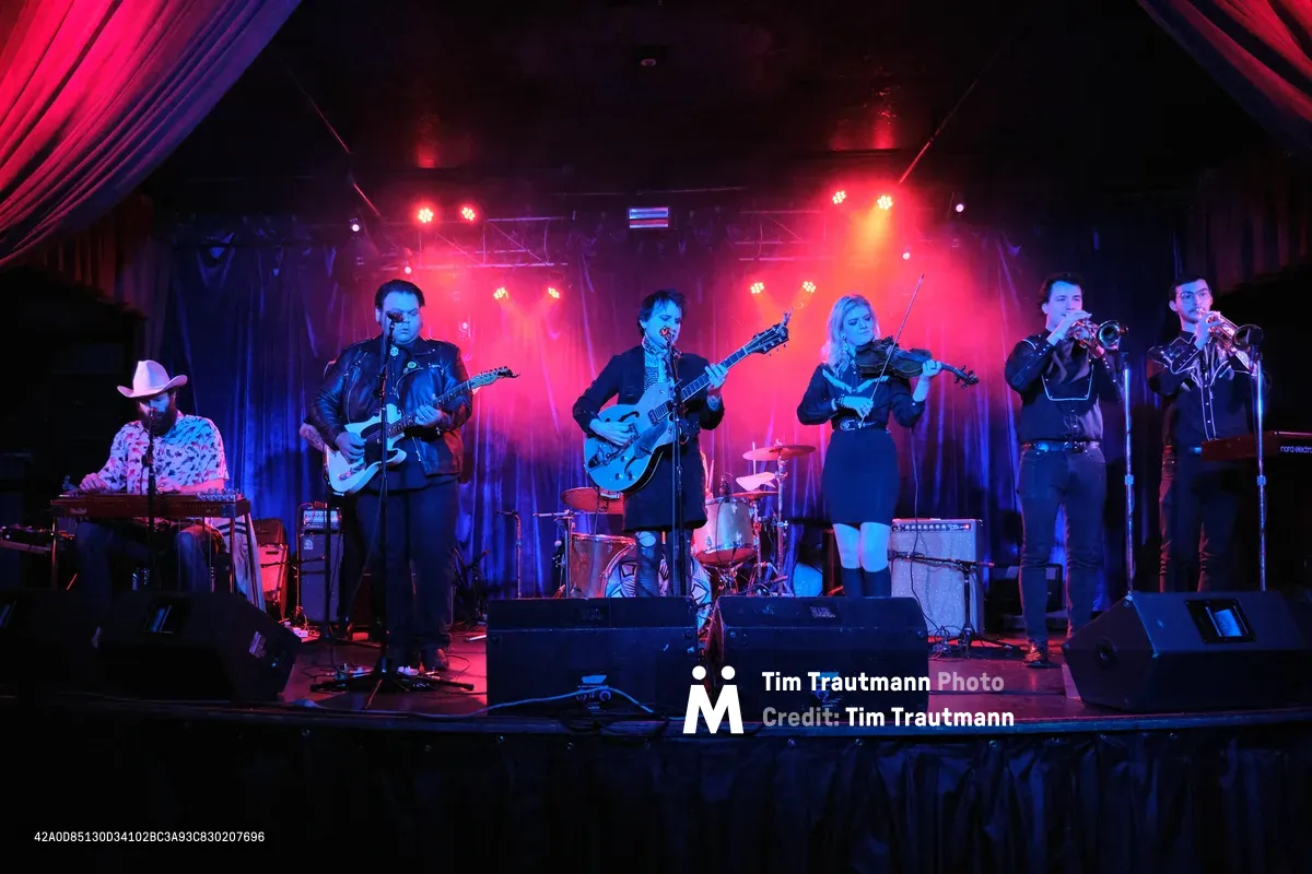Six musicians from Roselit Bone command the intimate stage at Dante's in Portland, bathed in saturated crimson and azure theatrical lighting. The ensemble features electric guitars, violin, brass instruments, and keyboards, creating a dynamic country-fusion performance. Dramatic spotlights pierce through the smoky venue atmosphere, casting the performers in theatrical silhouettes against draped curtains and exposed stage rigging.