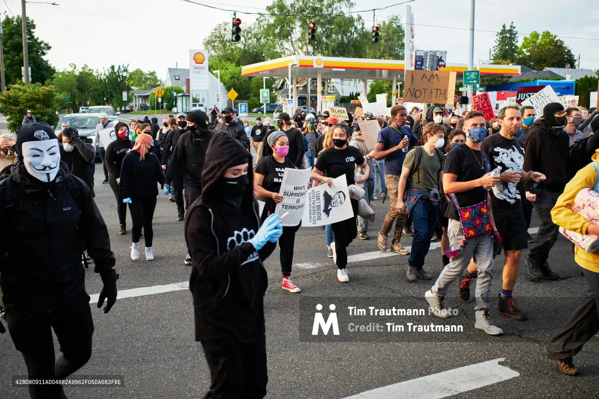 A diverse crowd of protesters moves through the intersection at Northeast 33rd Avenue in Portland's Grant Park neighborhood during a Black Lives Matter demonstration following George Floyd's murder. Participants wear face masks and carry handmade signs, with some donning Guy Fawkes masks as they march past a Shell gas station under overcast skies. The scene captures the raw energy and determination of community activism in suburban Portland, where residential homes and commercial establishments form the backdrop for this moment of social uprising.
