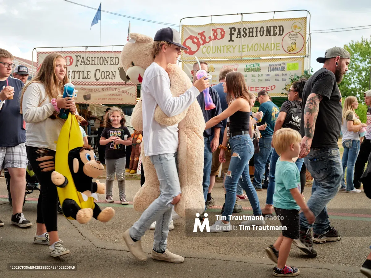 A bustling crowd of fairgoers walks through the Clark County Fair in Vancouver, Washington. In the center of the frame, a teenage boy in a white sweatshirt and black cap carries an enormous stuffed teddy bear he won at a carnival game. To his left, a girl carries a large stuffed monkey in a banana costume. Families, children, and adults fill the midway, with food vendor signs for Old Fashioned Country Lemonade and Old Fashioned Kettle Corn visible in the background. Several people carry colorful fair drinks.