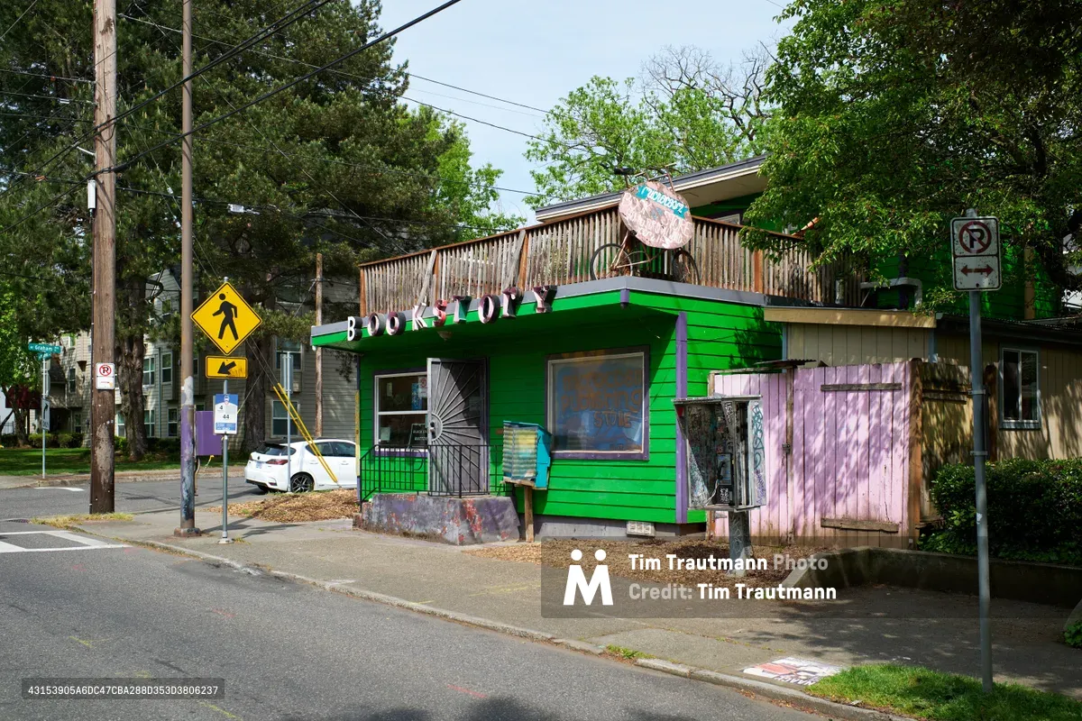 A whimsically painted independent bookstore anchors a tree-lined residential corner in Portland's historic Eliot district. The small building bursts with personality through its bold green and purple exterior, hand-painted signage, and eclectic architectural details that speak to the area's creative spirit. A public pay phone stands nearby, adding to the street's urban character. Dappled afternoon light filters through mature street trees, casting gentle shadows across the quiet intersection where urban infrastructure meets neighborhood charm.