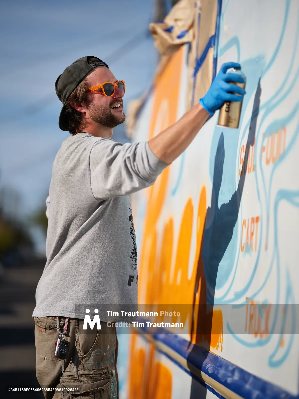 A bearded muralist in orange sunglasses and backwards cap applies bright blue paint to a colorful food truck exterior in Portland's King neighborhood. The artist works with focused concentration, his paint-stained cargo pants and blue latex gloves testament to hours of creative labor. Orange and blue hues dominate the emerging artwork against the soft afternoon sky, while the food truck's white canvas transforms under his skilled brushstrokes.