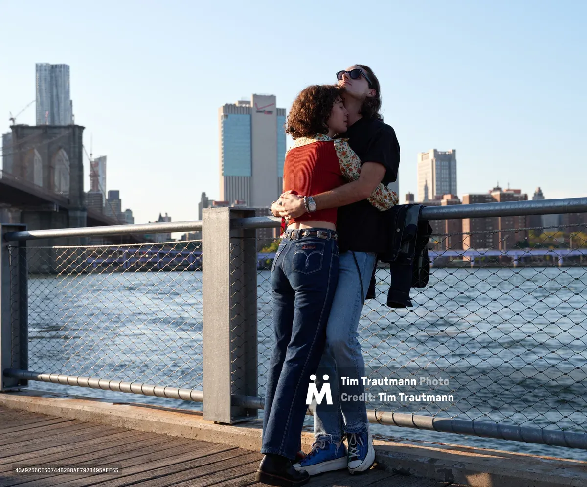 A young couple shares an intimate embrace on the DUMBO waterfront promenade, silhouetted against the golden hour light filtering across the East River. The woman in a vibrant orange ribbed sweater and the man in dark clothing stand pressed together at the chain-link fence, with the towering Manhattan skyline and Brooklyn Bridge creating a dramatic urban backdrop. The warm evening light bathes the wooden boardwalk and distant skyscrapers in honeyed tones, capturing a tender moment of connection amid the city's grandeur.