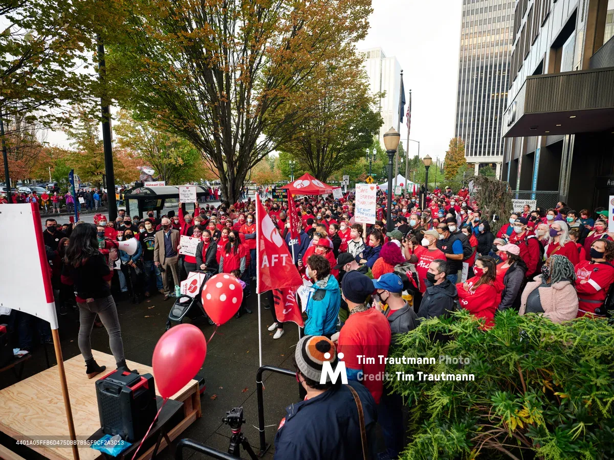 Hundreds of nurses and healthcare workers in vibrant red union attire congregate outside Kaiser Permanente's towering glass headquarters in Portland's Lloyd District, their determined faces reflecting the gravity of impending labor action. The autumn foliage of street trees creates a golden canopy above the crowd, while red AFT banners and protest signs punctuate the sea of solidarity. A makeshift speaking platform commands attention from the diverse assembly, capturing the pivotal moment before a significant healthcare strike unfolds against the urban backdrop of downtown Portland.