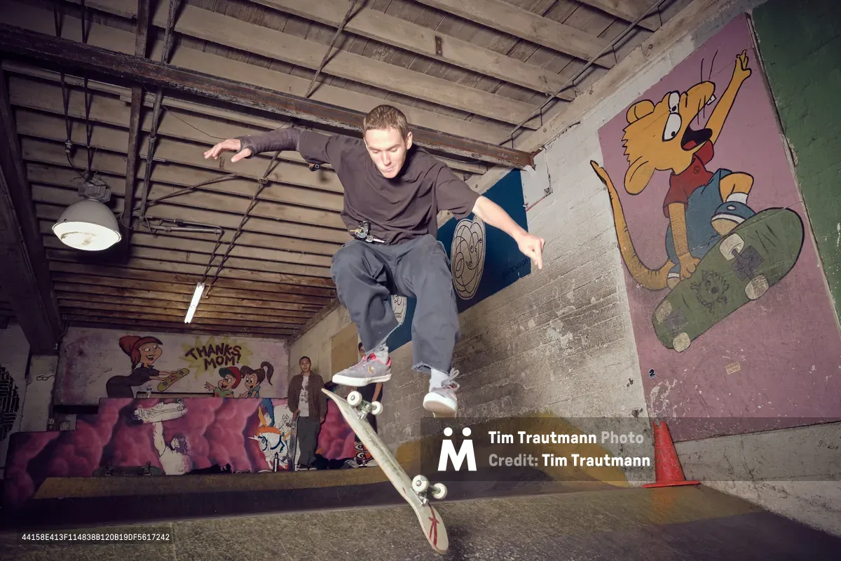 A skateboarder executes a dynamic aerial maneuver in the concrete bowl at Commonwealth Skateboarding in Portland's Buckman neighborhood. The exposed wooden beam ceiling and weathered industrial pendant lights frame the action, while vibrant cartoon murals featuring animated characters create a nostalgic backdrop. The rider, caught mid-flight in baggy street wear, embodies the raw energy of underground skate culture against the gritty urban canvas of this Southeast Portland institution.
