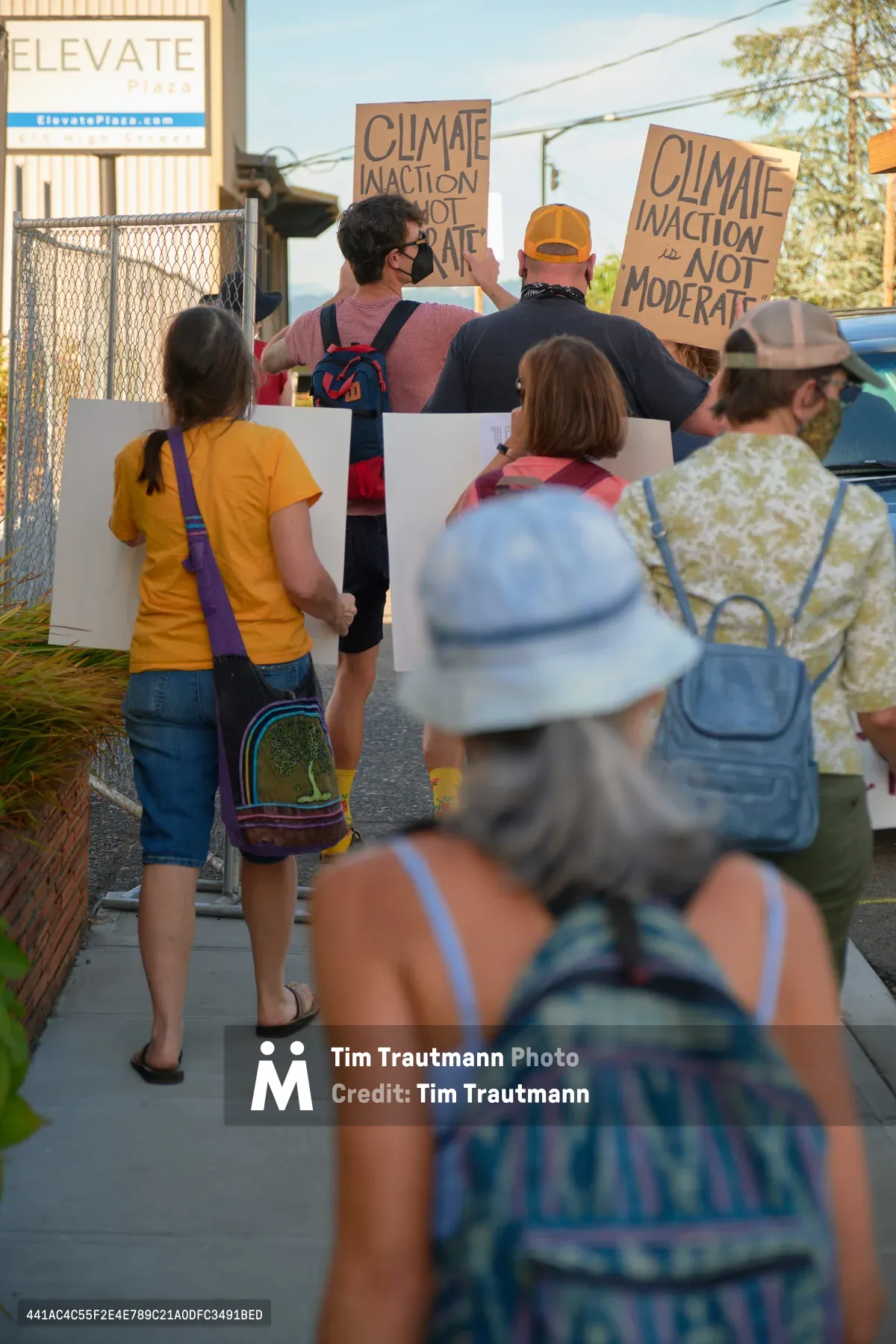 A determined group of climate activists gathers on the sidewalk outside Representative Kurt Schrader's office in Oregon City, their handwritten cardboard signs declaring "Climate Inaction Is Not Moderate." Golden afternoon light filters through the suburban setting as protesters of various ages stand unified along McLoughlin Promenade, some wearing masks and carrying colorful bags, creating an intimate yet powerful scene of grassroots political engagement. The composition captures both the urgency of their message and the everyday nature of democratic participation in small-town America.