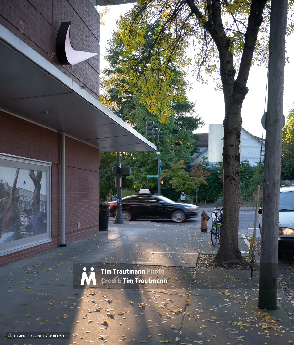 A dark sedan blurs past the Nike Factory Store along Northeast Knott Street in Portland's Eliot neighborhood during the golden hour of an autumn evening. Fallen leaves carpet the sidewalk beneath mature street trees, while traffic lights cast a gentle glow over the retail streetscape. The scene captures the quiet rhythm of urban life in this historic Albina district, where the prominent Nike outlet meets neighborhood tranquility.