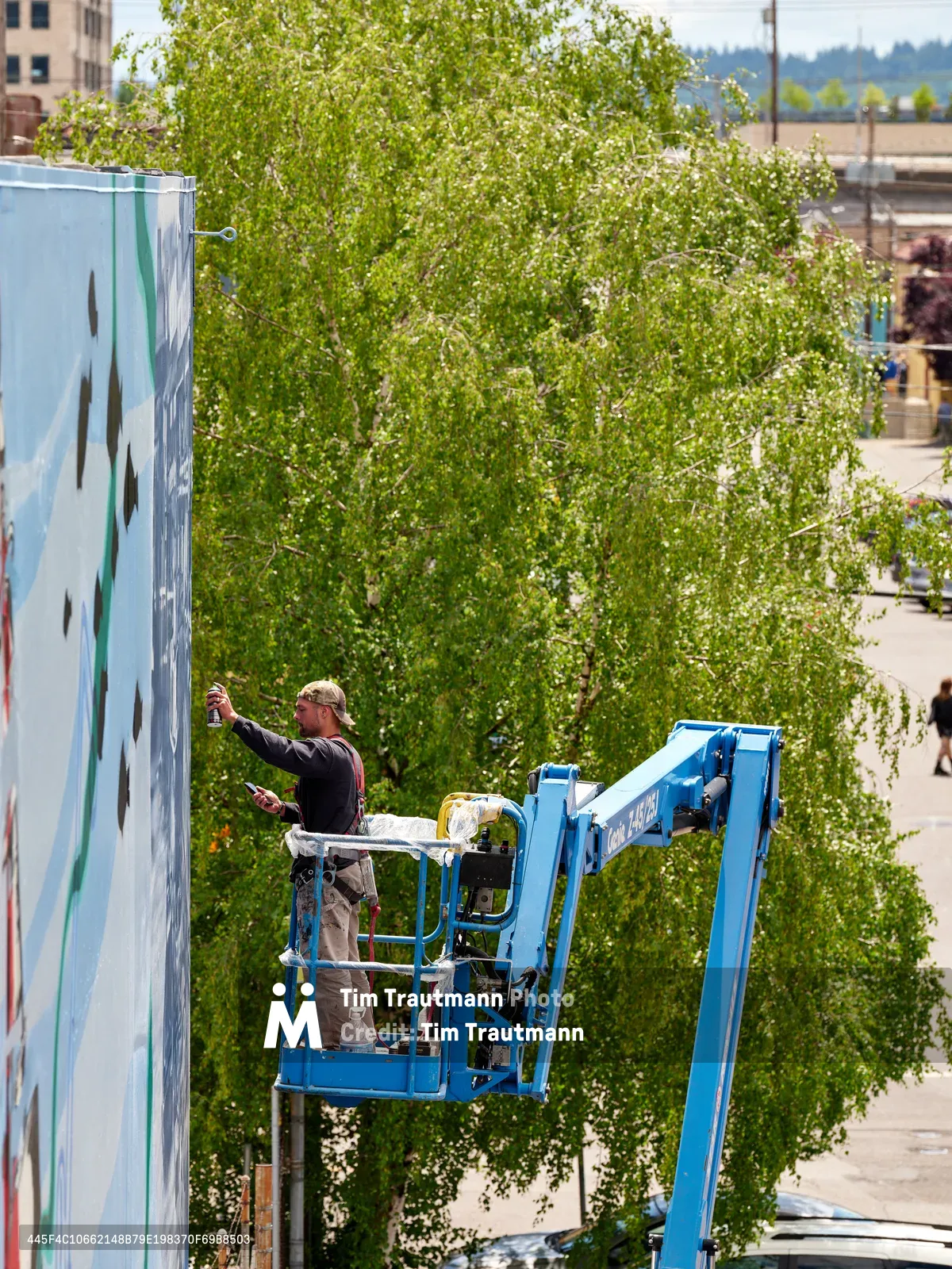 Two artists, Jeremy Nichols and Devin Finley, work from a blue aerial lift platform to create a large-scale mural on the side of a building in Southeast Portland. The scene captures them mid-creation against a backdrop of spring foliage from a mature tree, with urban brick architecture visible in the frame. Bright midday sunlight illuminates their concentrated work on the Portland Street Art Alliance project, highlighting the intersection of public art and community transformation in Portland's Central Eastside district.