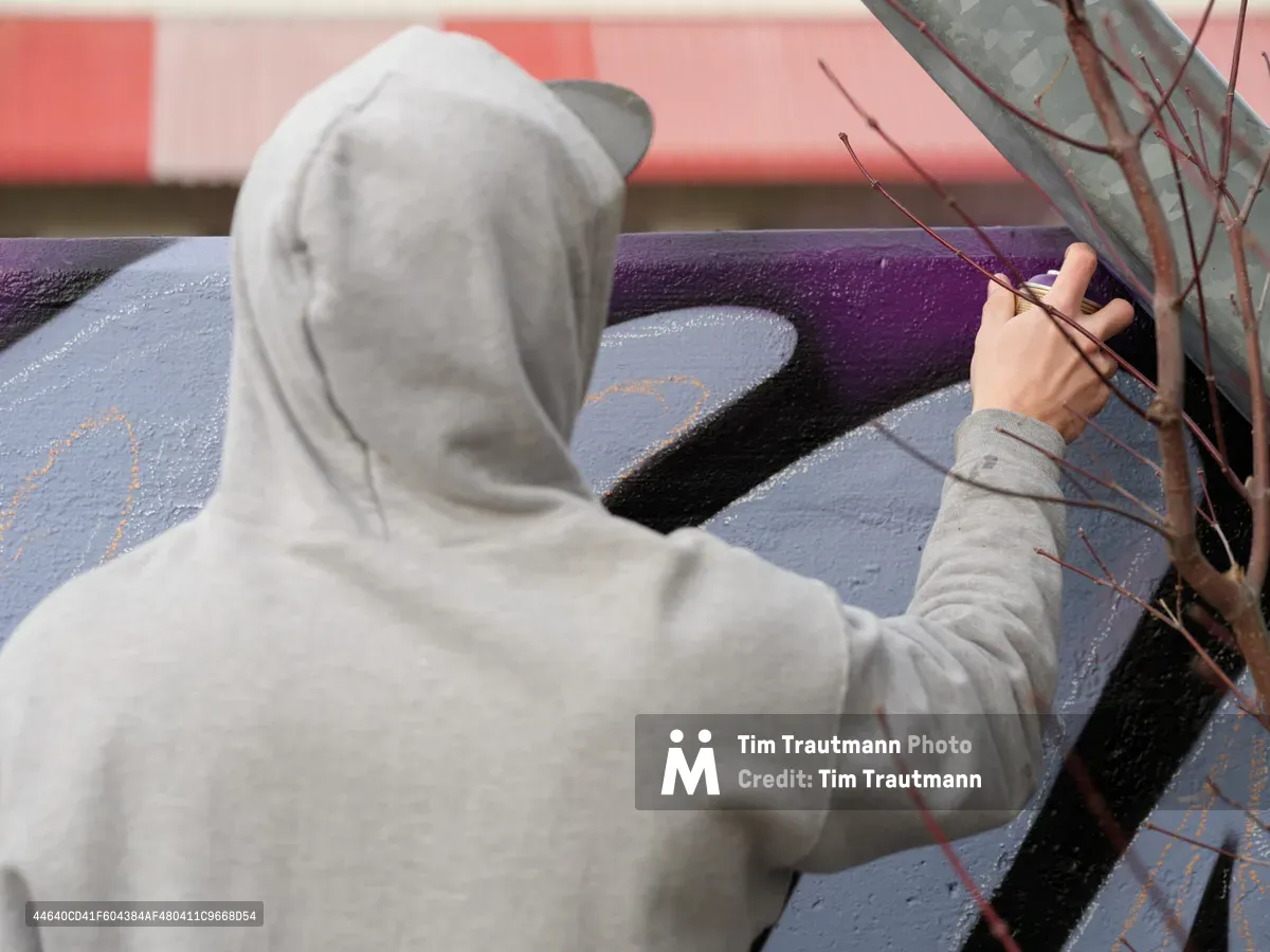 Travis Fields, known as Campographic, works intently on a vibrant mural at the Clay Creative Building in Portland's Hosford-Abernethy neighborhood. Wearing a gray hoodie, the artist reaches toward his canvas of purple, blue, and black abstract forms painted on weathered concrete. The intimate perspective captures the focused concentration of street art creation, with dried branches adding organic texture to the urban creative process.