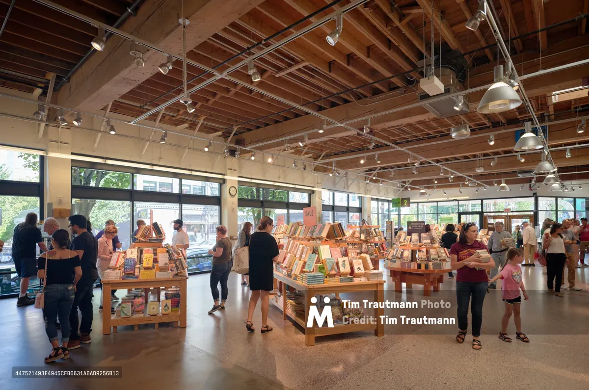 Inside Powell's City of Books in Portland's Pearl District, warm afternoon light streams through expansive windows, illuminating browsing customers amid carefully arranged wooden display tables. The industrial-chic ceiling reveals exposed beams and track lighting above the bustling literary sanctuary, where families and book lovers of all ages navigate between organized displays of featured titles. The polished concrete floors reflect the gentle glow of the space, creating an inviting atmosphere that captures the essence of Portland's renowned independent bookstore culture.