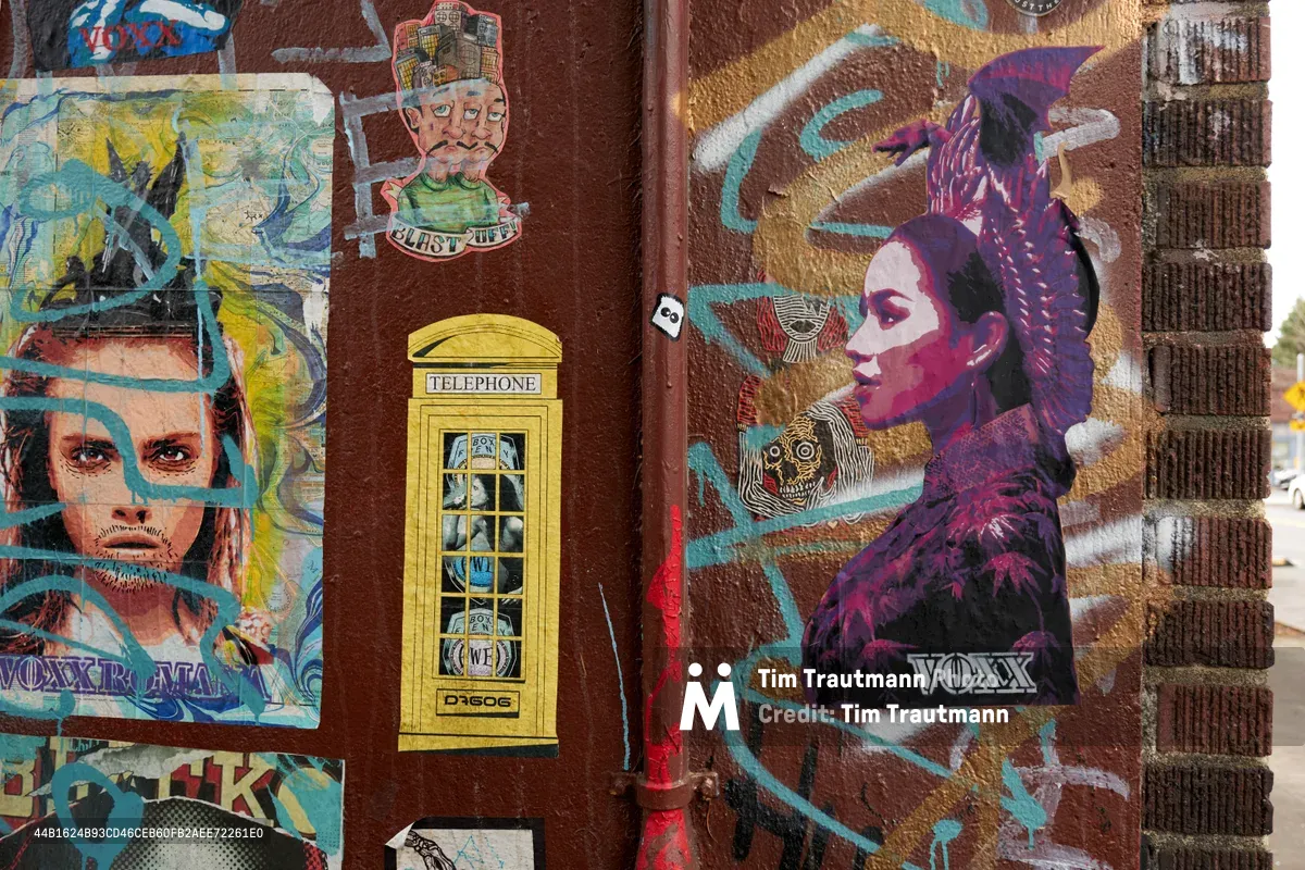 A vibrant street art installation adorns a weathered brick wall in Portland's Concordia neighborhood, featuring layered portraits and pop culture imagery. A miniature yellow telephone booth serves as the focal point among collaged faces rendered in electric blues, purples, and turquoise. The mixed-media composition includes vintage-style character illustrations and contemporary portraiture, creating a dense urban tapestry that speaks to the eclectic creative spirit of Northeast Portland's artistic community.
